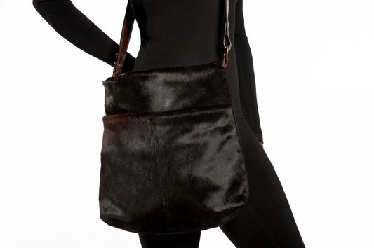 A dark brown cowhide leather messenger bag from SWABY Chicago Boutique, worn crossbody by a person in a black outfit. The bag measures 19 inches tall by 17 inches wide, featuring a rich, textured leather surface with two exterior slide-in pockets and a short leather handle. A long, adjustable strap is visible, complementing the bag’s sleek, minimalist design against a plain background.