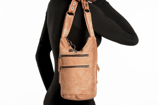 Small camel suede bucket bag with leather buckle straps worn as shoulder bag, featuring compact silhouette and multiple exterior zip pockets against black outfit.