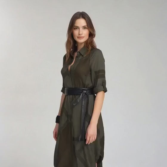Load video: A video of the Shernett No. 7 Shirt Dress in army green. Midi-length silhouette with collared button placket, rolled elbow-length sleeves with tab closures, diagonal tonal paneling, asymmetric draped hemline, and black leather obi wrap belt. By SWABY - A New York Fashion Designer.