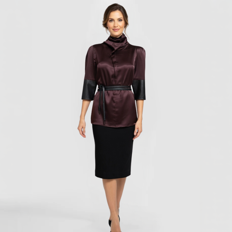 A fashion model poses in the Roya w/s Dress, by Chicago Fashion Designer SWABY. The Roya Dress is a sophisticated burgundy satin turtleneck top with draped cowl collar, subtle puff shoulders, contrasting black faux-leather three-quarter sleeves, and black tie belt cinching the waist, styled over a black pencil skirt for a polished, professional look.