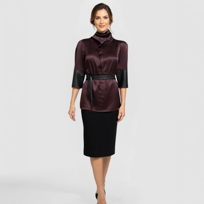 A fashion model poses in the Roya w/s Dress, by Chicago Fashion Designer SWABY. The Roya Dress is a sophisticated burgundy satin turtleneck top with draped cowl collar, subtle puff shoulders, contrasting black faux-leather three-quarter sleeves, and black tie belt cinching the waist, styled over a black pencil skirt for a polished, professional look.